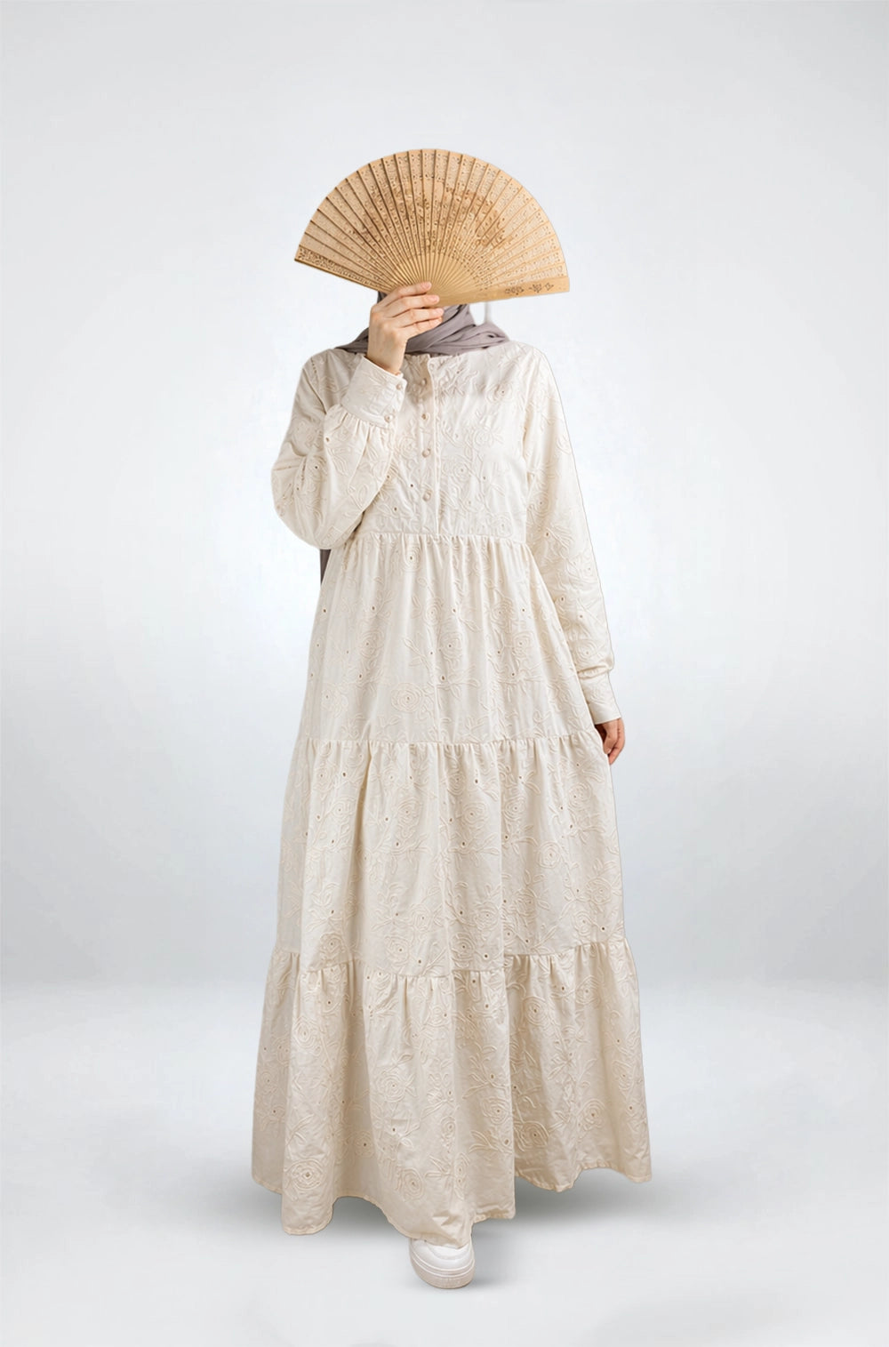 Person wearing a long beige dress holding a wooden fan on a white background